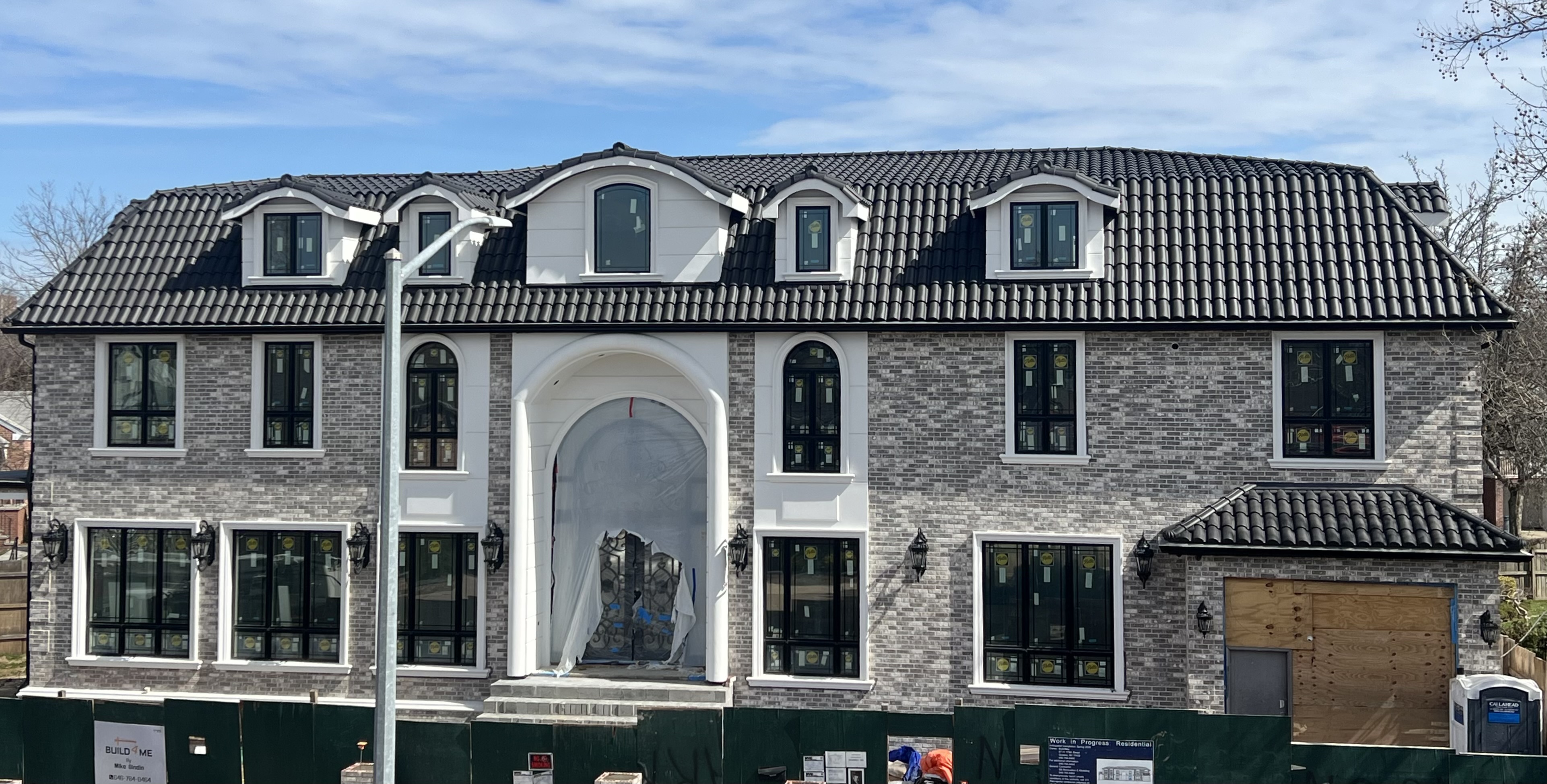 Luxury gray brick mansion under construction with dark charcoal slate S-tile clay roof, arched windows, white trim, and multiple dormers